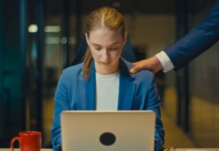Young woman in a blue blazer working late at the office, looking down at her laptop. A male colleague in a suit places a hand on her shoulder. Concept of workplace abuse and power dynamics. By HudHudPro - AdobeStock