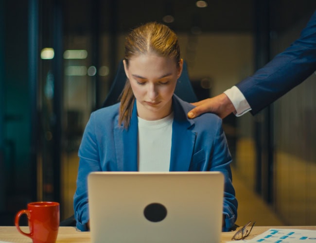 Young woman in a blue blazer working late at the office, looking down at her laptop. A male colleague in a suit places a hand on her shoulder. Concept of workplace abuse and power dynamics. By HudHudPro - AdobeStock