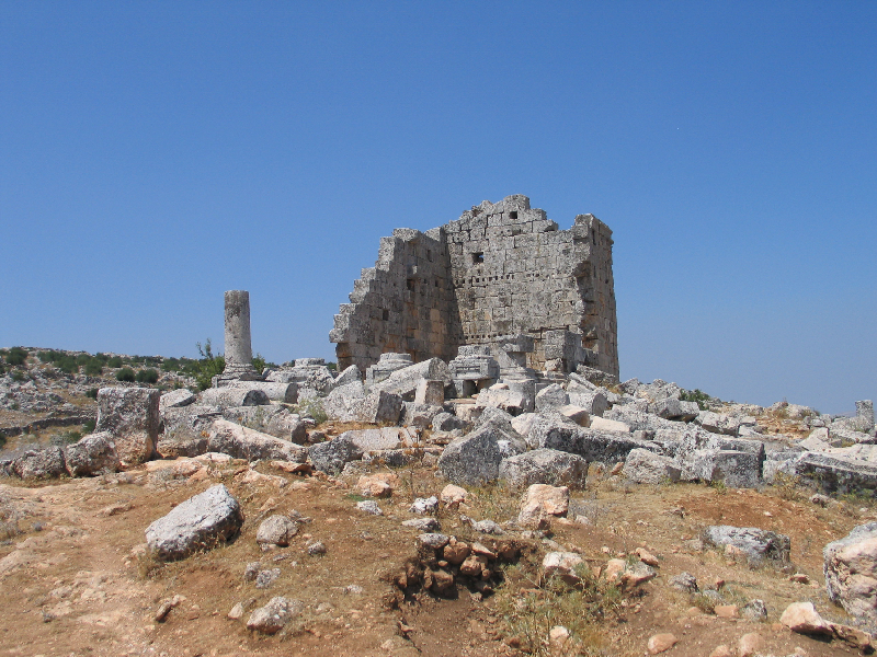 ruine d'un temple composé d'un mur droit avec deux angles éboulés, des pierres taillées pêle-mêle au sol, une colonne étêtée est dressée à gauche du mur