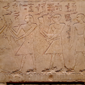 bas relief égyptien, 4 personnages sont représenté en pied, celle de gauche fait face aux trois autres. La pierre semble être un linteau. Des inscriptions en hiéroglyphe sont gravé au dessus des personnages