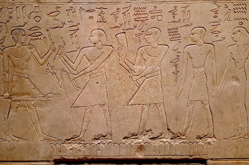 bas relief égyptien, 4 personnages sont représenté en pied, celle de gauche fait face aux trois autres. La pierre semble être un linteau. Des inscriptions en hiéroglyphe sont gravé au dessus des personnages